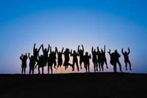 dark blue sky with silhouettes of hill and 18 people jumping in the air with hands up.