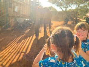 two little girls; one looking and smiling at the camera and the back of ones head looking at elephants walking towards them.