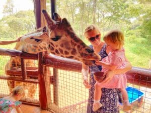 mother holding daughter while daughter feeds a giraffe from her hand.