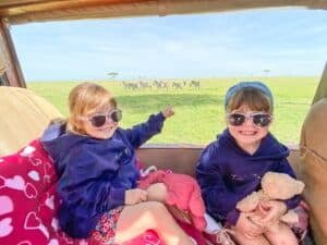 two little girls wearing matching purple hoodies and sunglasses smiling with zebras in a field behind them in the distance.