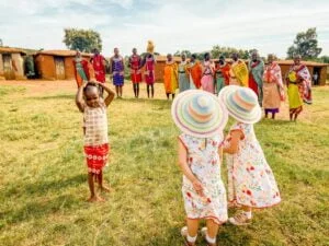Two little girls wearing matching white dresses with flowers and sun hats look at Kenya tribe standing in front of their village.