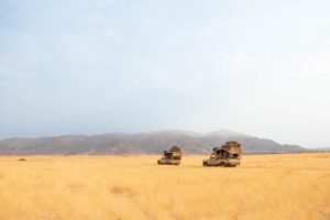 Field with mountains in the background and two large jeeps driving with equipment on top.