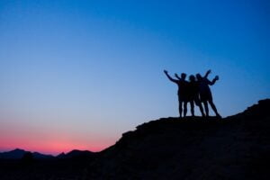 Blue and pink sky showing silhouettes of a cliff with three people standing with arms in the air.