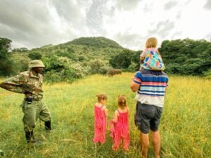 Tour guide in army patterned clothing standing with arms on hips while father with toddler on shoulders and two girls wearing matching pink jumpsuits look out into a field at a rhino.