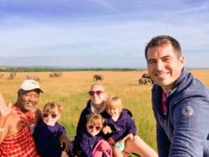 Family and tour guide smiling on vehicle in front of a field full of elephants.